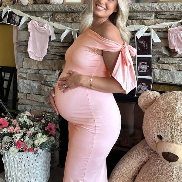 Roze zwangerschaps maxi jurk met strik, one-shoulder en elegante pasvorm, ideaal voor een babyshower.